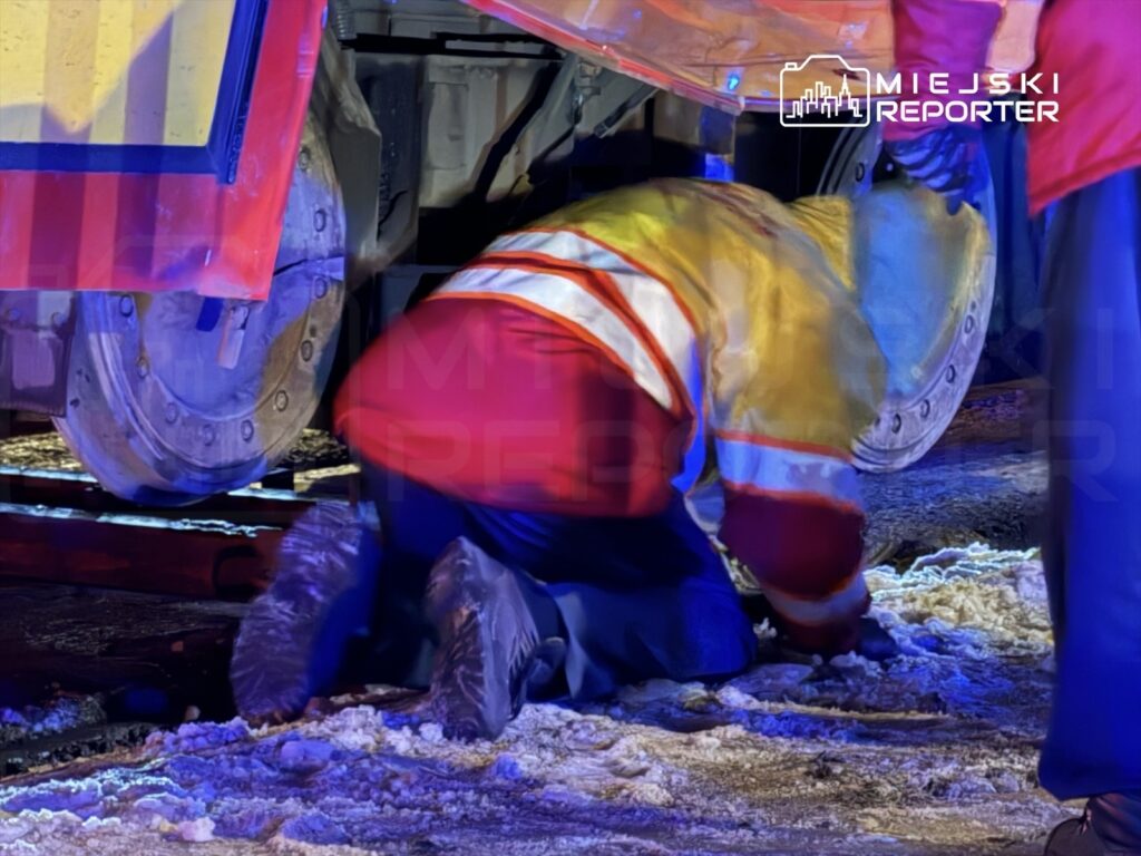 Pracownik w odblaskowej kamizelce klęczy przy pojeździe, sprawdzając jego podwozie w oświetlonym, zimowym otoczeniu.