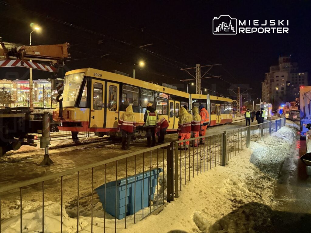 Grupa pracowników w odblaskowych kamizelkach prowadzi prace przy tramwaju na oświetlonym torowisku w zimowej scenerii.