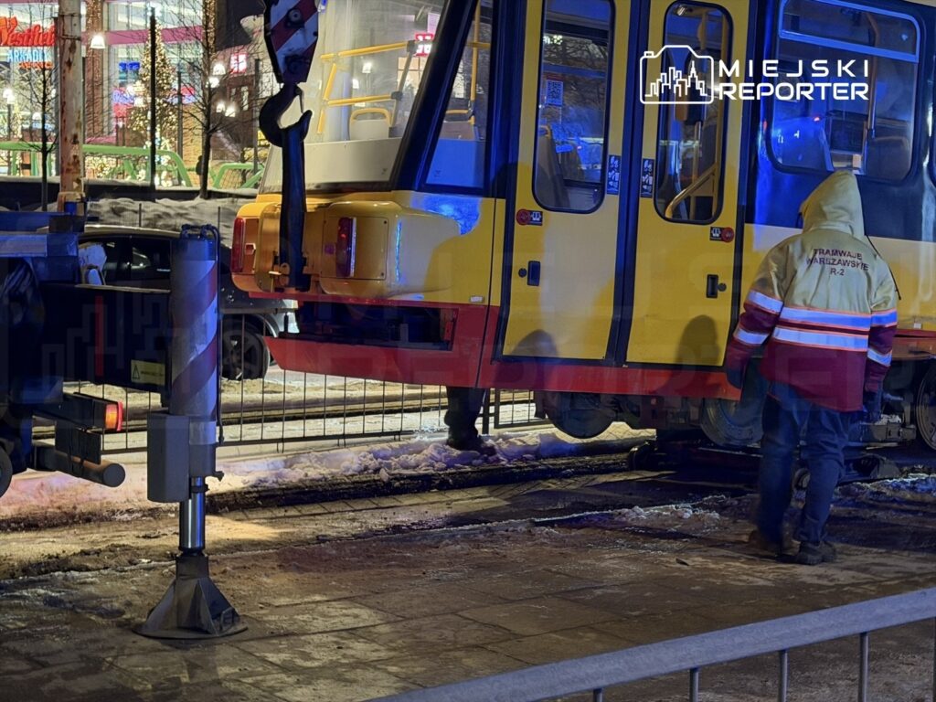 Pracownik w odblaskowej kurtce nadzoruje podnoszenie tramwaju za pomocą dźwigu na zaśnieżonej ulicy.