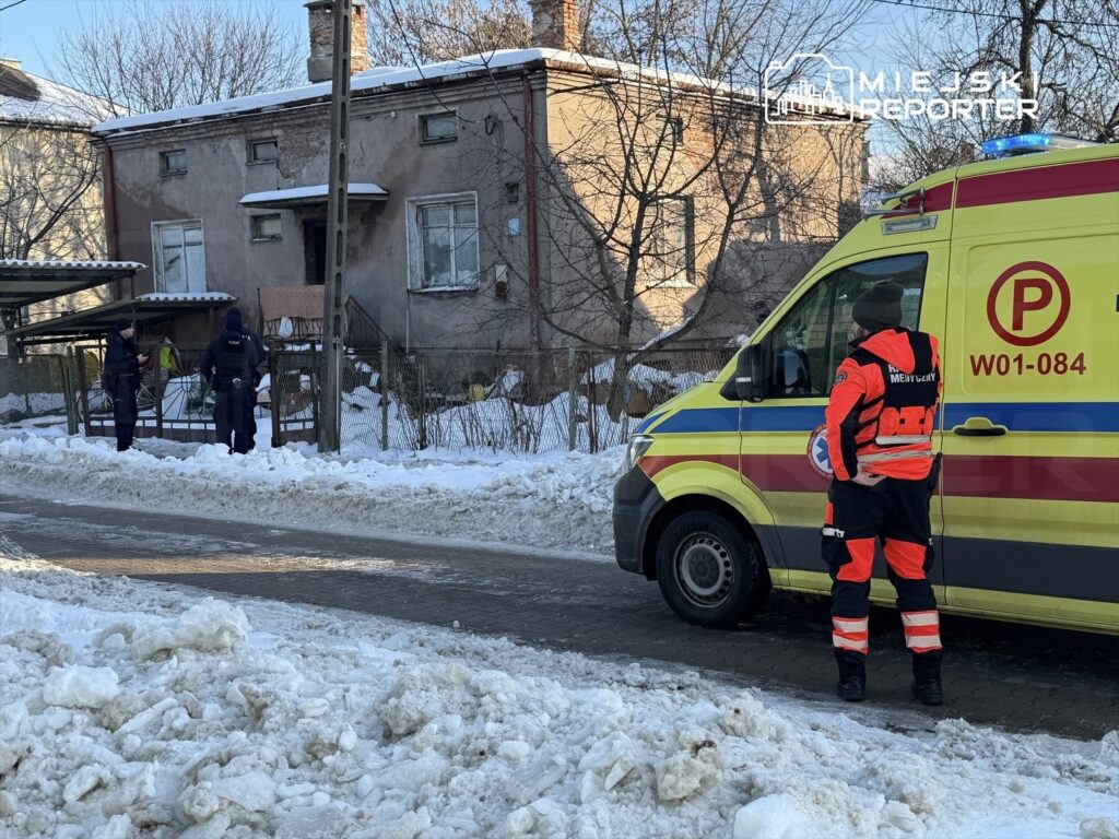 Funkcjonariusze policji rozmawiają przy wejściu do zaniedbanego budynku, obok stoi karetka w zimowym otoczeniu.