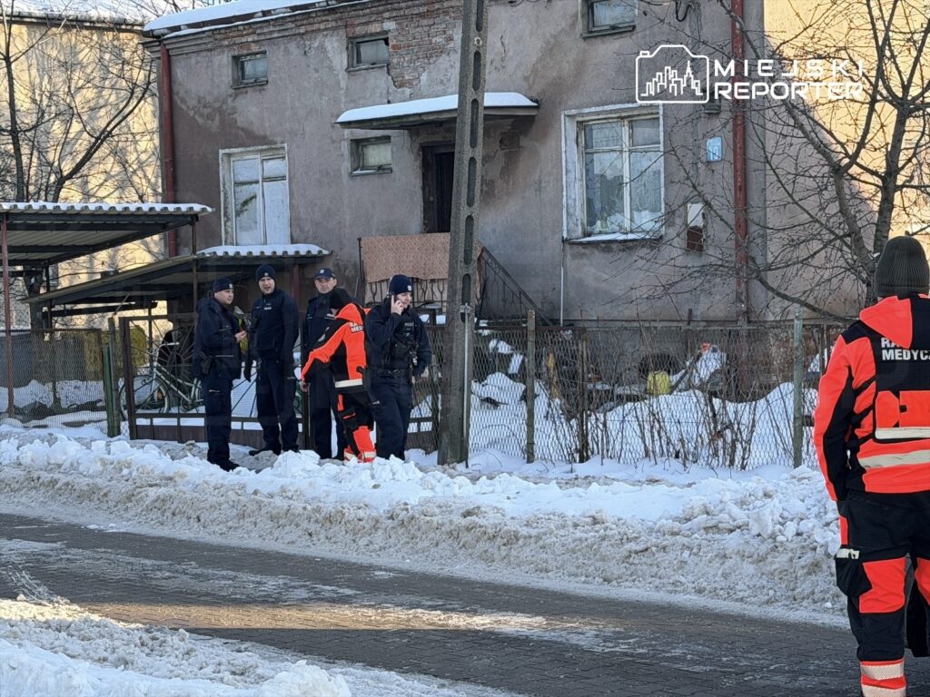 Grupa pięciu funkcjonariuszy w mundurach rozmawia na chodniku przed starym budynkiem, w otoczeniu śniegu.