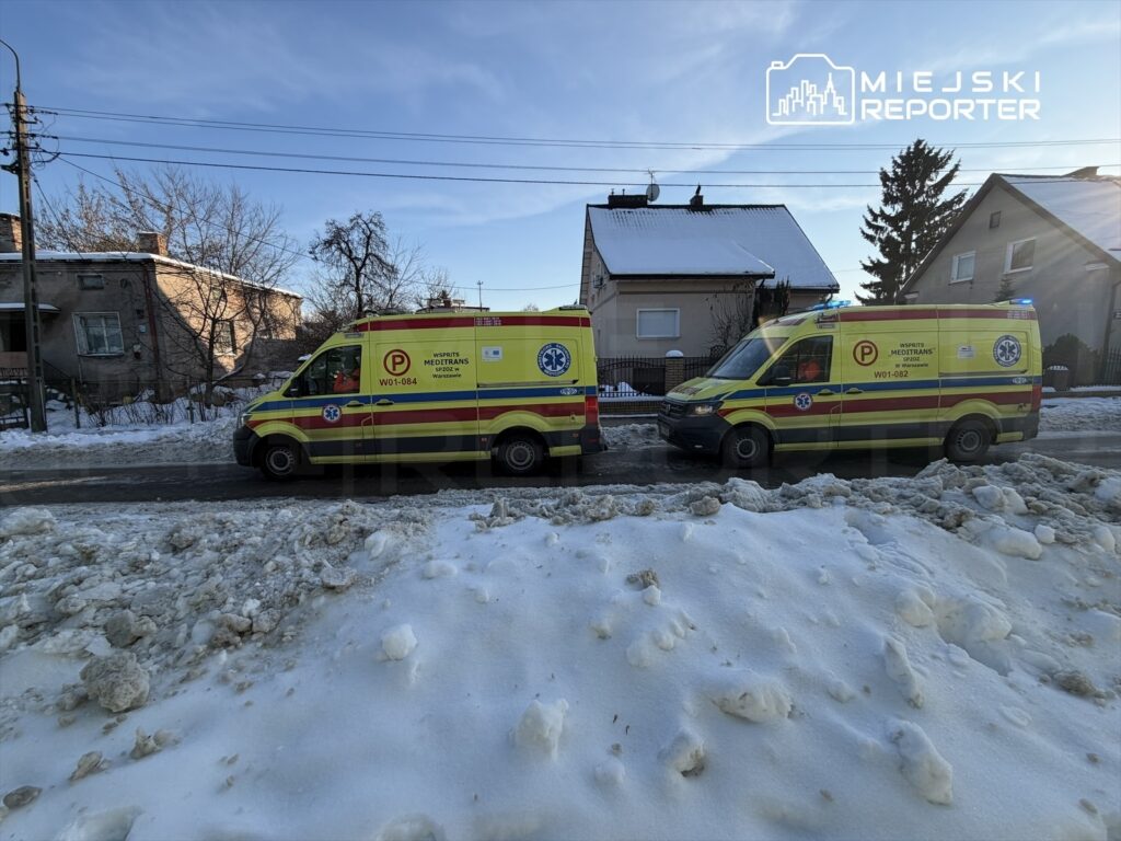 Dwa karetki pogotowia stoją na ośnieżonej drodze obok budynku mieszkalnego, z włączonymi sygnalizatorami.