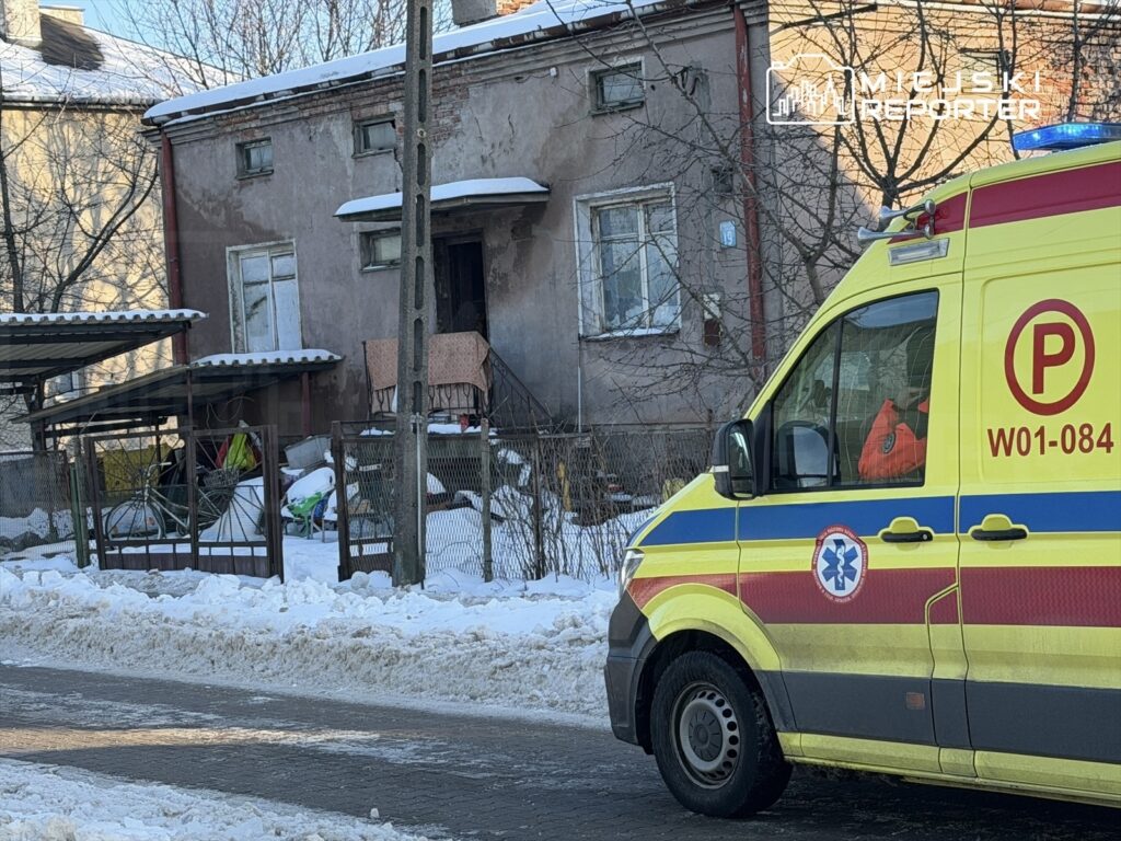 Karetka pogotowia stojąca na zaśnieżonej ulicy obok budynku mieszkalnego z widocznym balkonem.