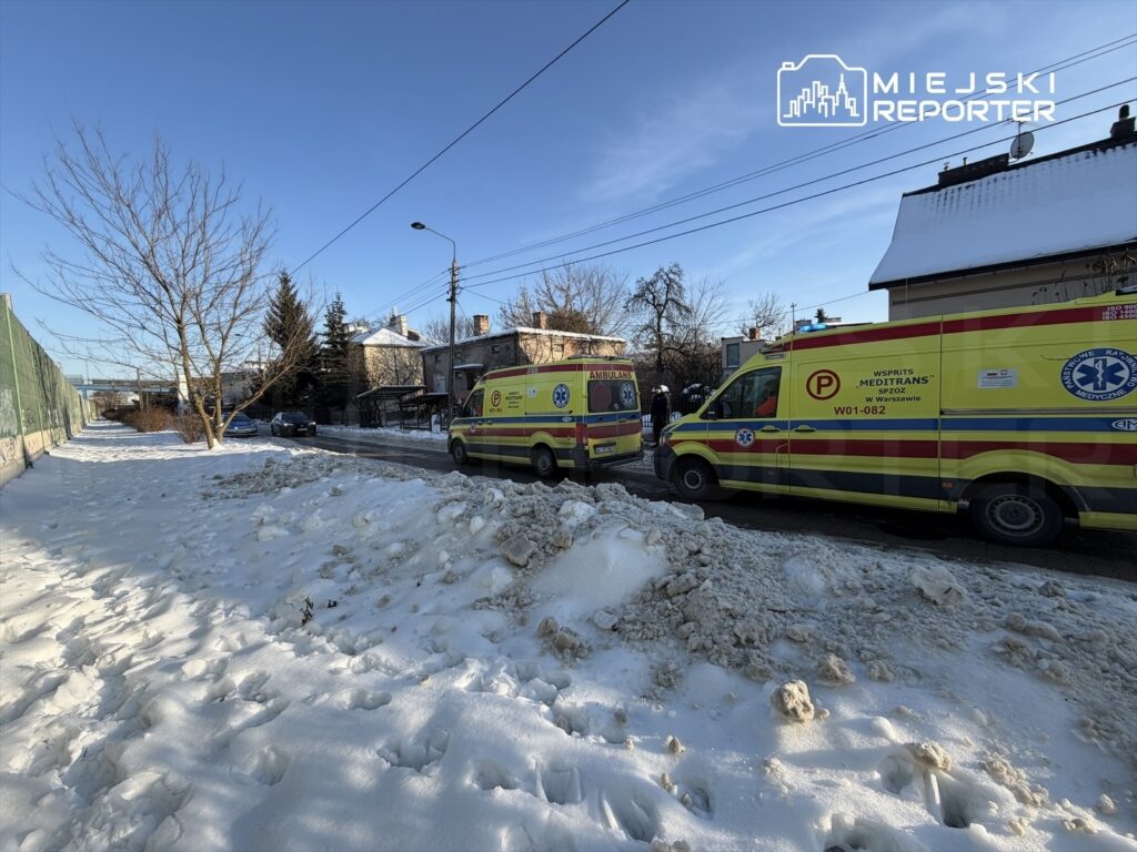 Dwa karetki pogotowia zaparkowane na zaśnieżonej ulicy, obok domów jednorodzinnych i drzew.