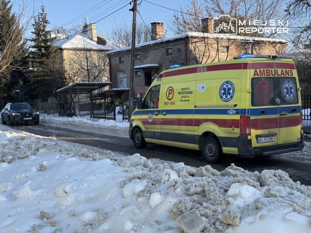 Ambulans z włączonymi sygnałami świetlnymi zatrzymany na zaśnieżonej ulicy obok czarnego samochodu osobowego.