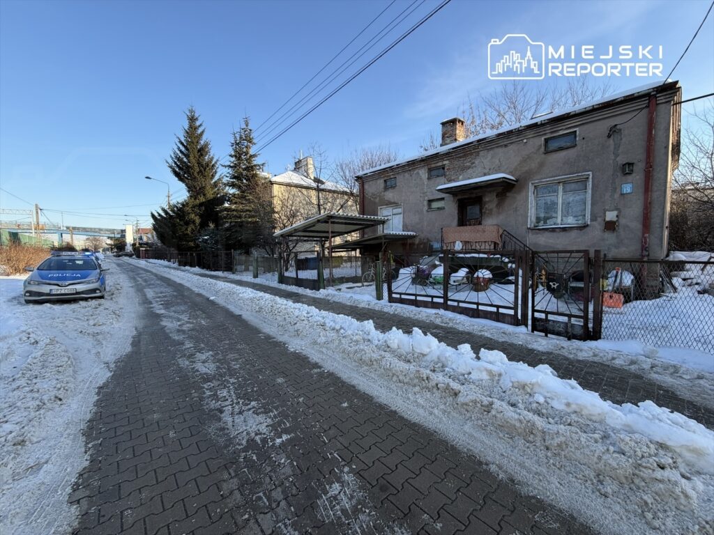 Policyjny radiowóz zaparkowany na zaśnieżonej ulicy obok starego, szarego budynku mieszkalnego.