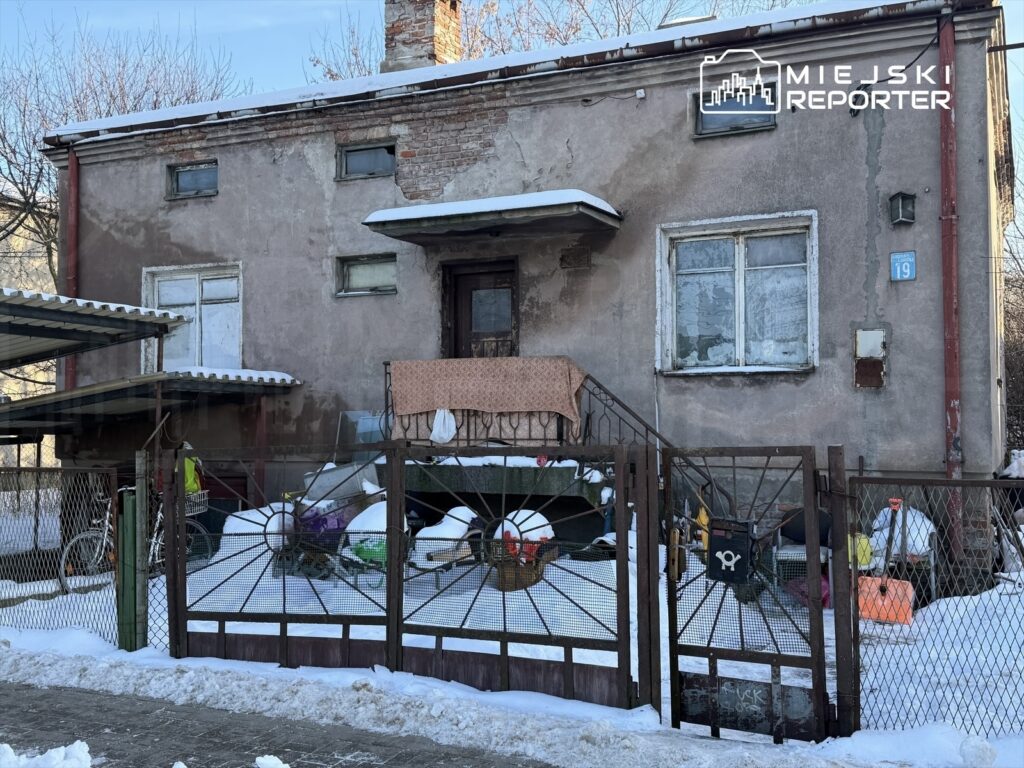 Zniszczony budynek mieszkalny z brudnymi oknami, otoczony śniegiem i starą siatką ogrodzeniową. Na podwórku widać porozrzucane przedmioty.