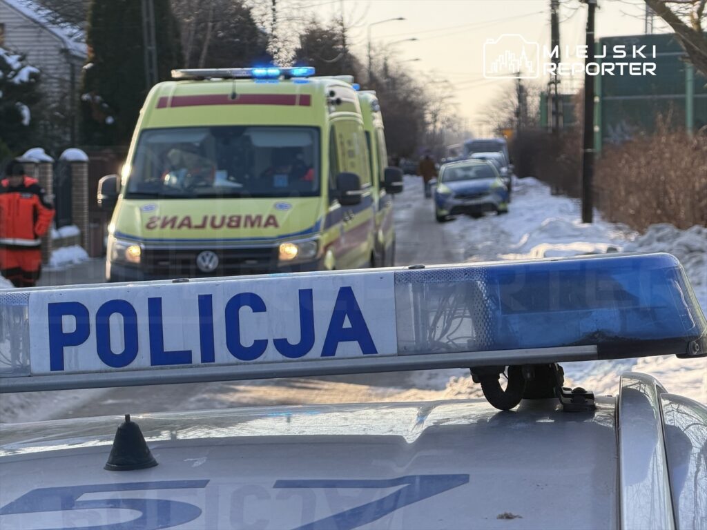 Radiowóz policyjny z napisem "POLICJA" na dachu stoi na zaśnieżonej ulicy, obok widać ambulans i ratowników medycznych.