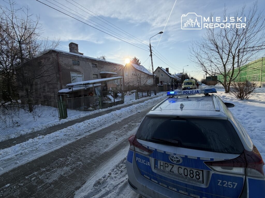 Policyjny radiowóz z włączonymi sygnałami świetlnymi stoi na zaśnieżonej ulicy obok starego budynku mieszkalnego.