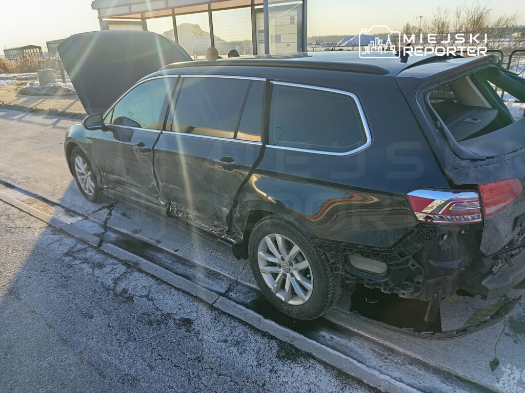 Uszkodzony czarny samochód osobowy z otwartą maską stoi na poboczu drogi w słoneczny dzień.
