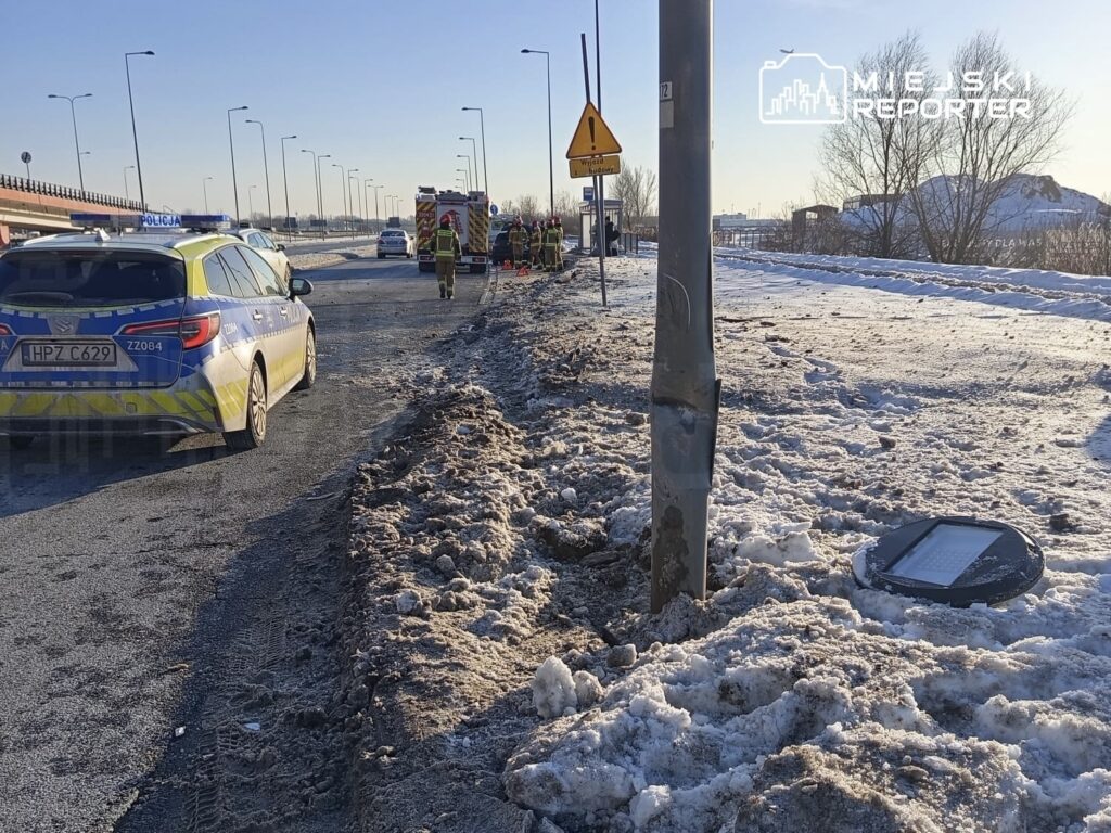 Na drodze przy zaspach śnieżnych stoi policyjny radiowóz, a obok pracują strażacy i ratownicy medyczni.