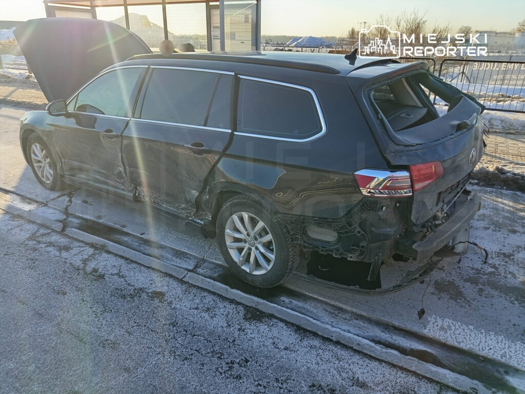 Uszkodzony czarny samochód osobowy z otwartą maską stoi na zaśnieżonej drodze, z widocznymi zarysowaniami na boku.