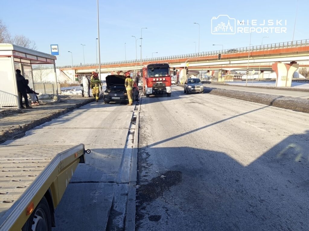 Dwóch strażaków sprawdza silnik samochodu osobowego na drodze, obok stoi wóz strażacki, a w tle widać przystanek autobusowy.