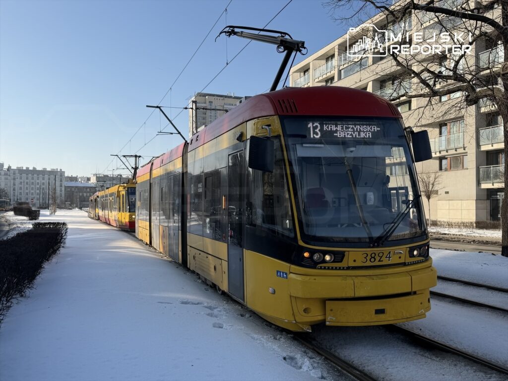Tramwaj linii 13 zatrzymany na przystanku, obok pokrytej śniegiem jezdni i nowoczesnych budynków mieszkalnych.