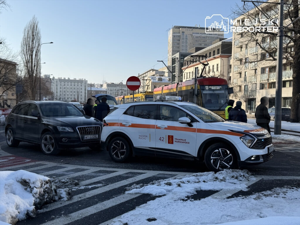 Czarny samochód osobowy stoi zderzony z białym pojazdem służb miejskich na zaśnieżonej ulicy. W tle widoczni przechodnie oraz tramwaj.