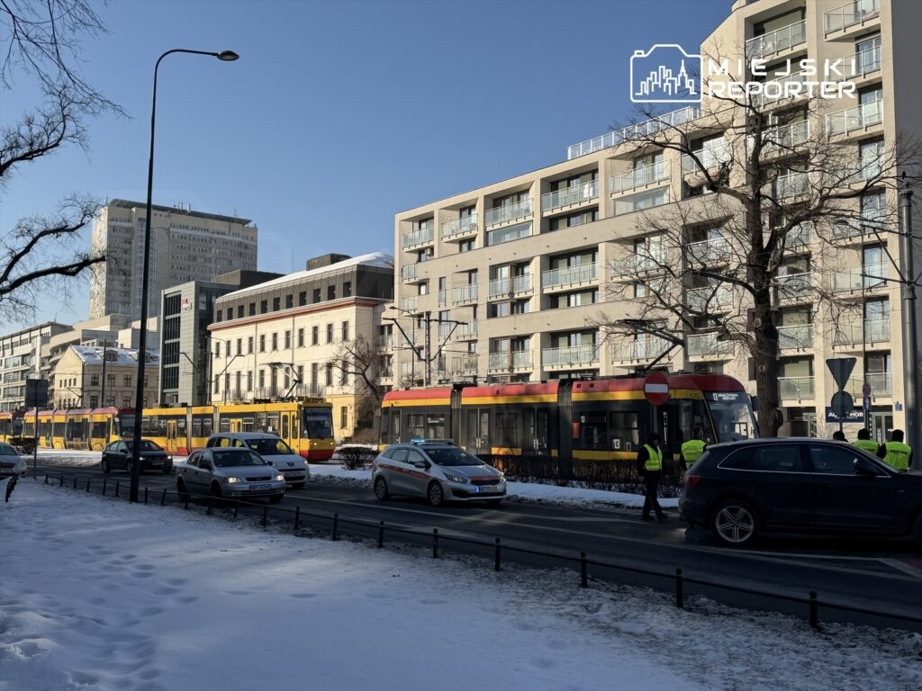 Tramwaj w kolorze żółto-czerwonym przejeżdża obok nowoczesnych budynków przy śnieżnej ulicy, gdzie stoją policjanci.