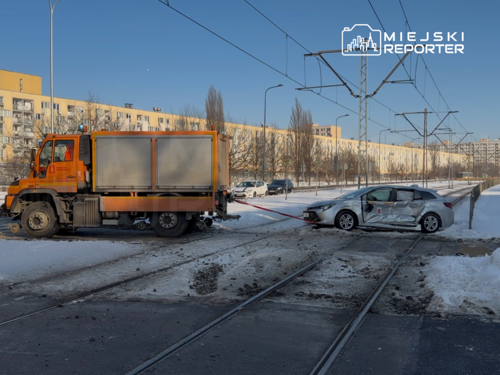 Ciężarówka z lawetą holuje uszkodzony samochód osobowy na torowisku w zimowej scenerii.