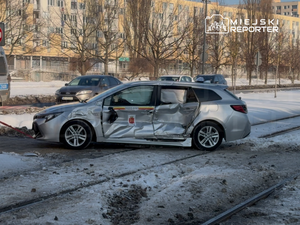 Uszkodzony srebrny samochód osobowy stoi na śniegu obok torów tramwajowych, z otwartymi drzwiami z boku.