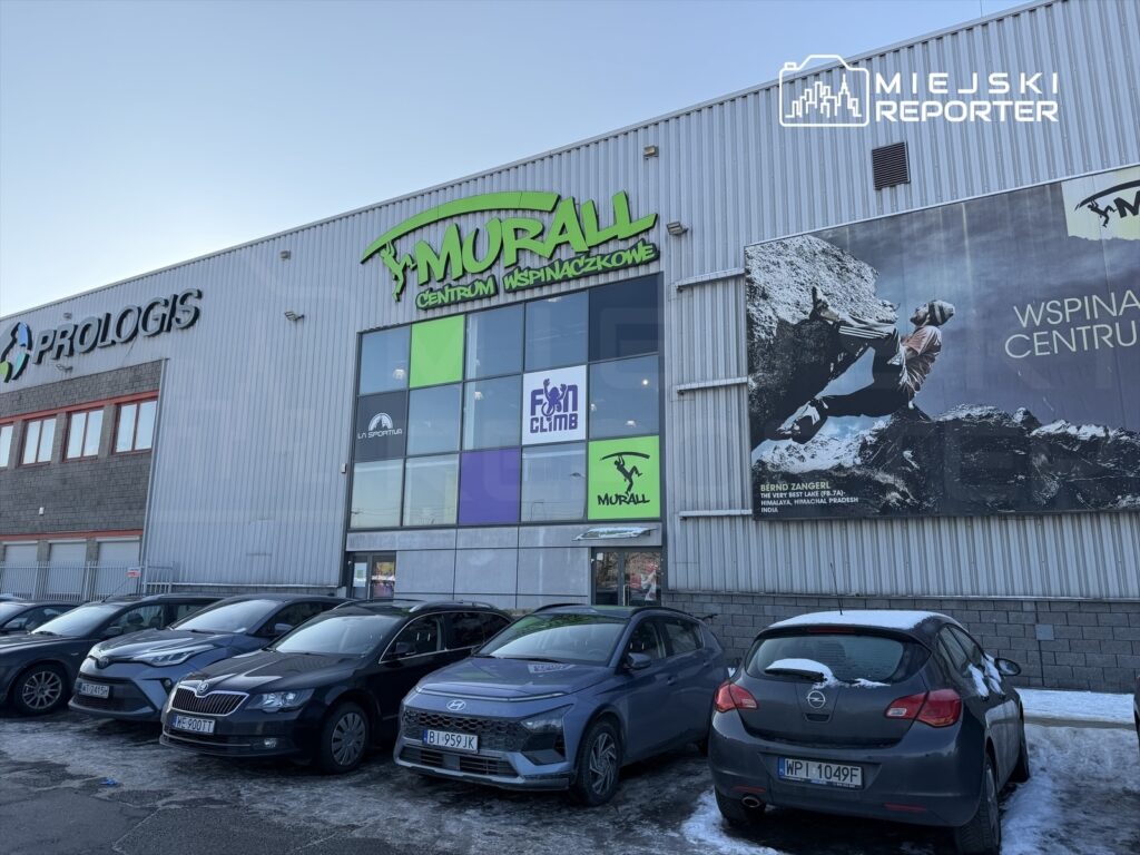 Centrum wspinaczkowe Murall z dużymi, kolorowymi znakami oraz reklamą wspinacza na ścianie budynku, przed którym zaparkowane są samochody.