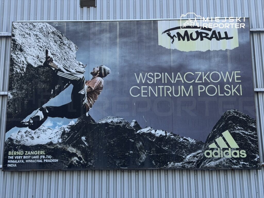 Mężczyzna wspinający się na skalną ścianę na tle górskiego krajobrazu, z reklamą "Wspinaczkowe Centrum Polski".