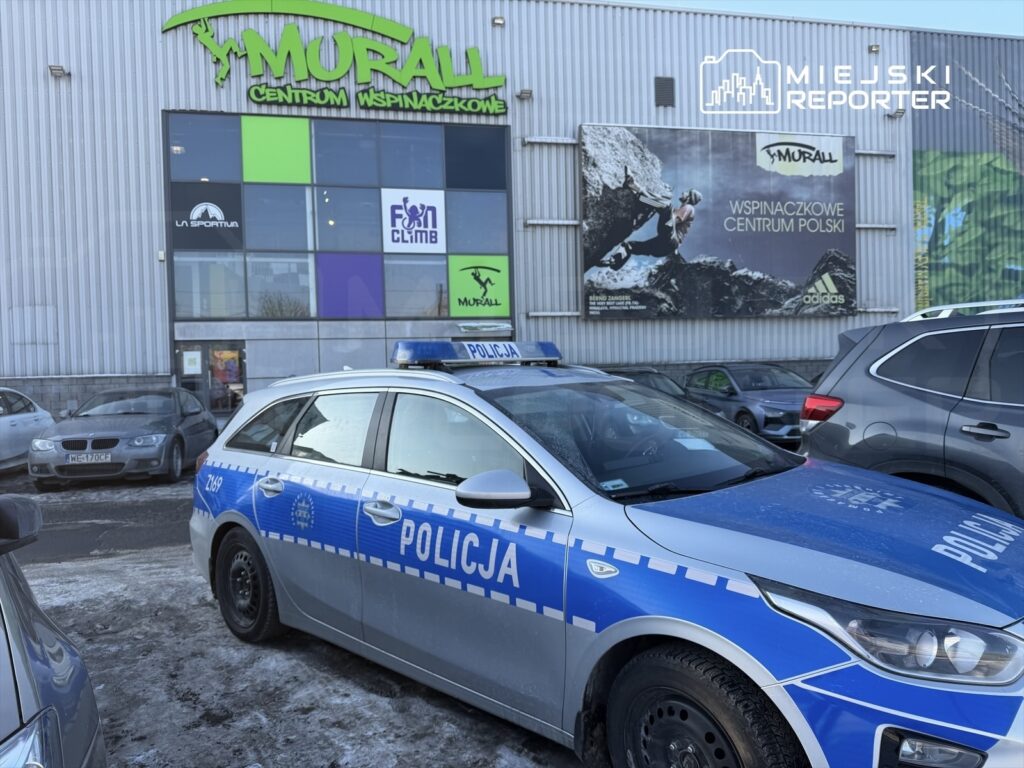 Policyjny radiowóz zaparkowany przed centrum wspinaczkowym z dużym banerem reklamowym.