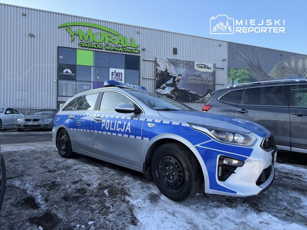 Policyjny samochód zaparkowany na parkingu przed centrum wspinaczkowym, obok innych pojazdów.