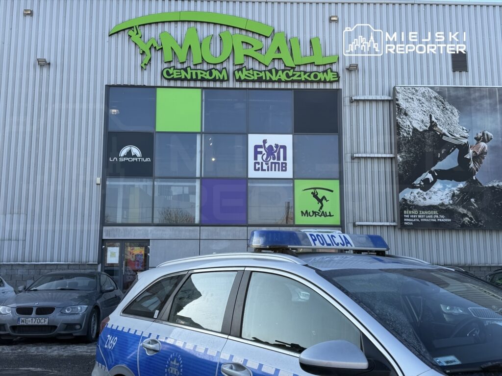 Centrum wspinaczkowe z dużymi witrynami i reklamą, obok zaparkowany radiowóz policji.