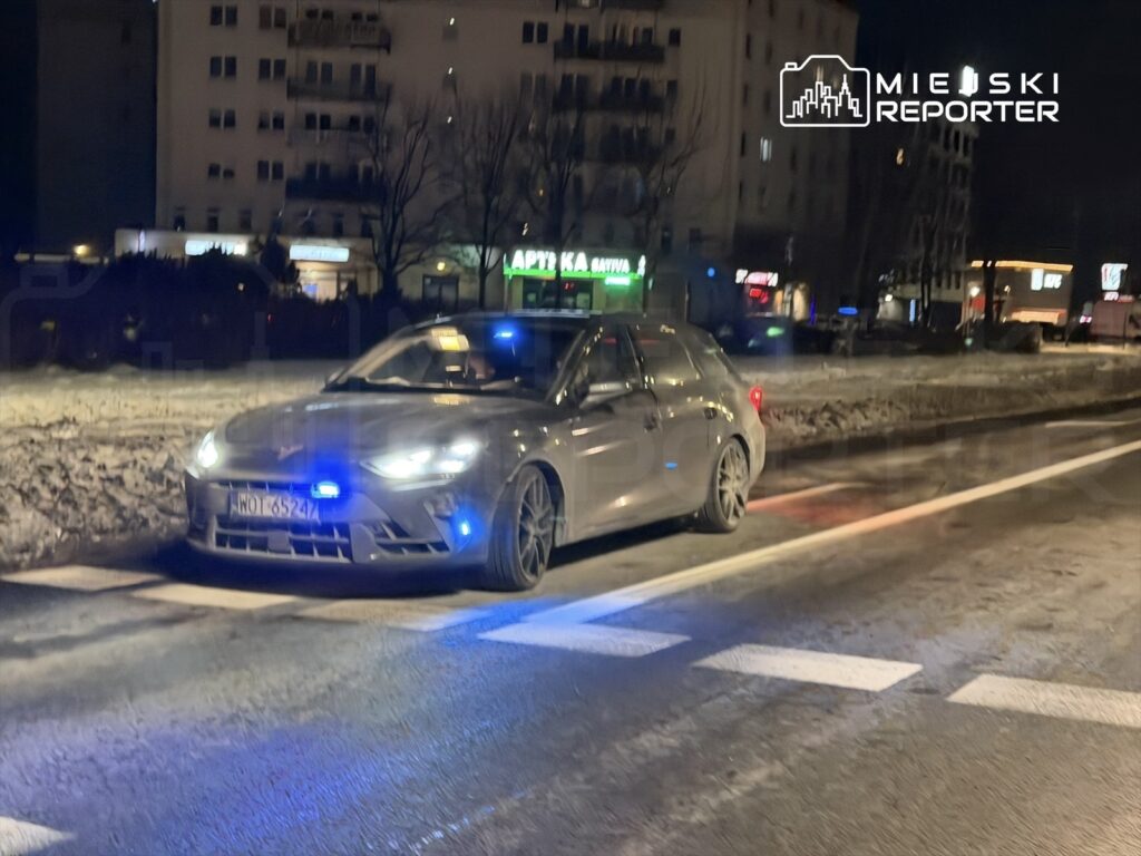 Radiowóz policyjny z włączonymi sygnalizatorami świetlnymi stoi na ulicy w nocy, obok oświetlonych budynków.