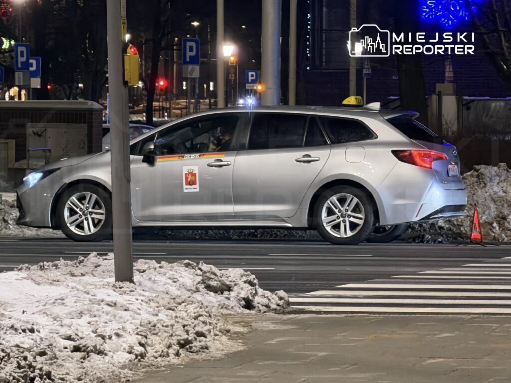 Taksi zaparkowane przy krawężniku na ulicy, obok śnieżnego pobocza, z włączonymi światłami.