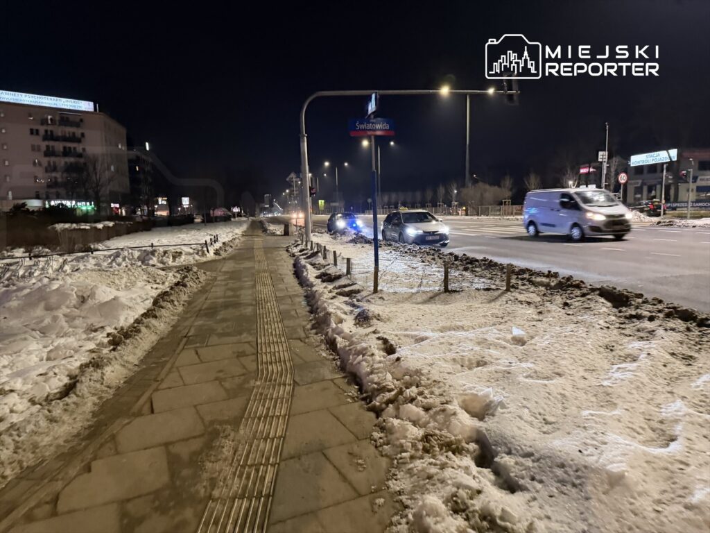Oświetlona ulica w nocy z widocznym chodnikiem, śniegiem oraz samochodami poruszającymi się po jezdni.