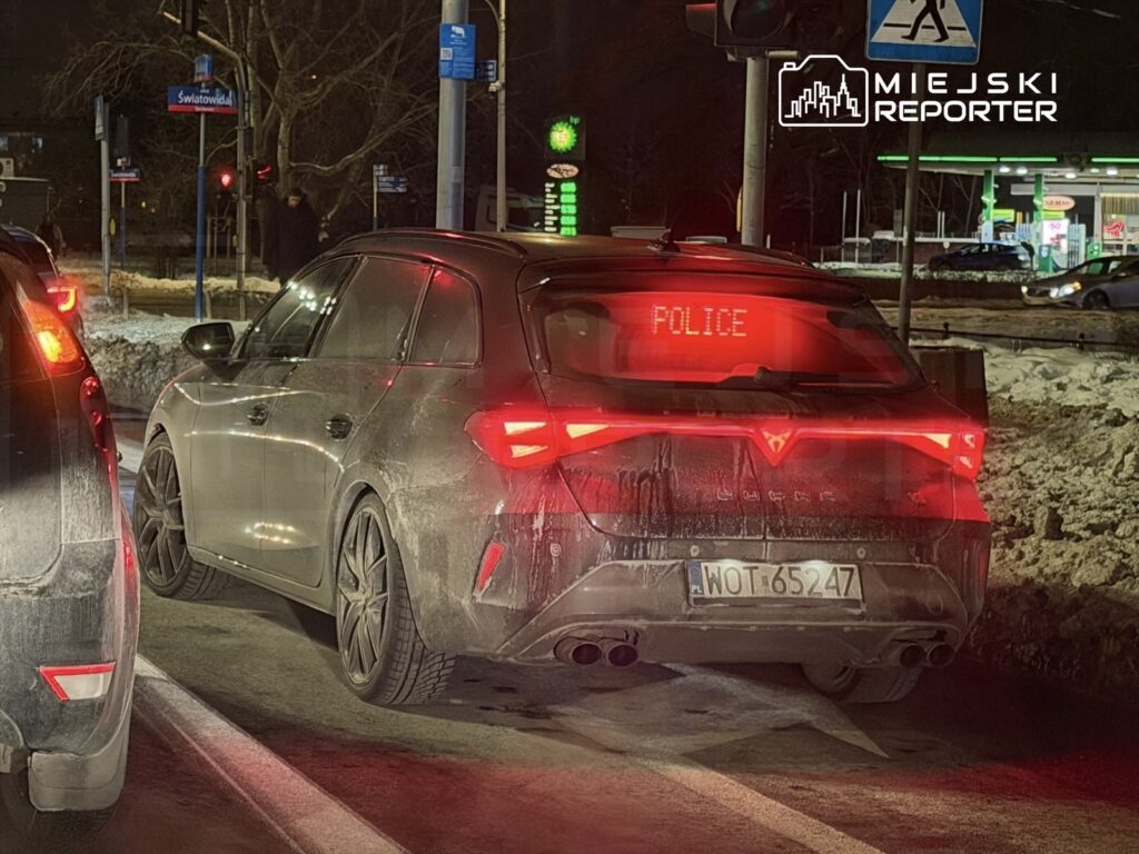 Policyjny samochód z widocznym napisem "POLICE" zatrzymany na ulicy w nocy, obok innych pojazdów.