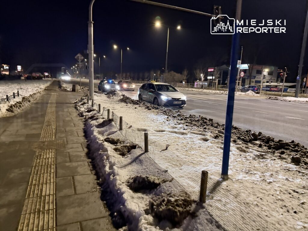 Samochody zaparkowane wzdłuż oświetlonej ulicy, obok chodnika pokrytego śniegiem i błotem.