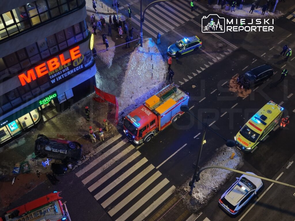 Na skrzyżowaniu ulicy leży przewrócone auto, obok niego strażacy i karetki, a w tle widoczny sklep meblowy.