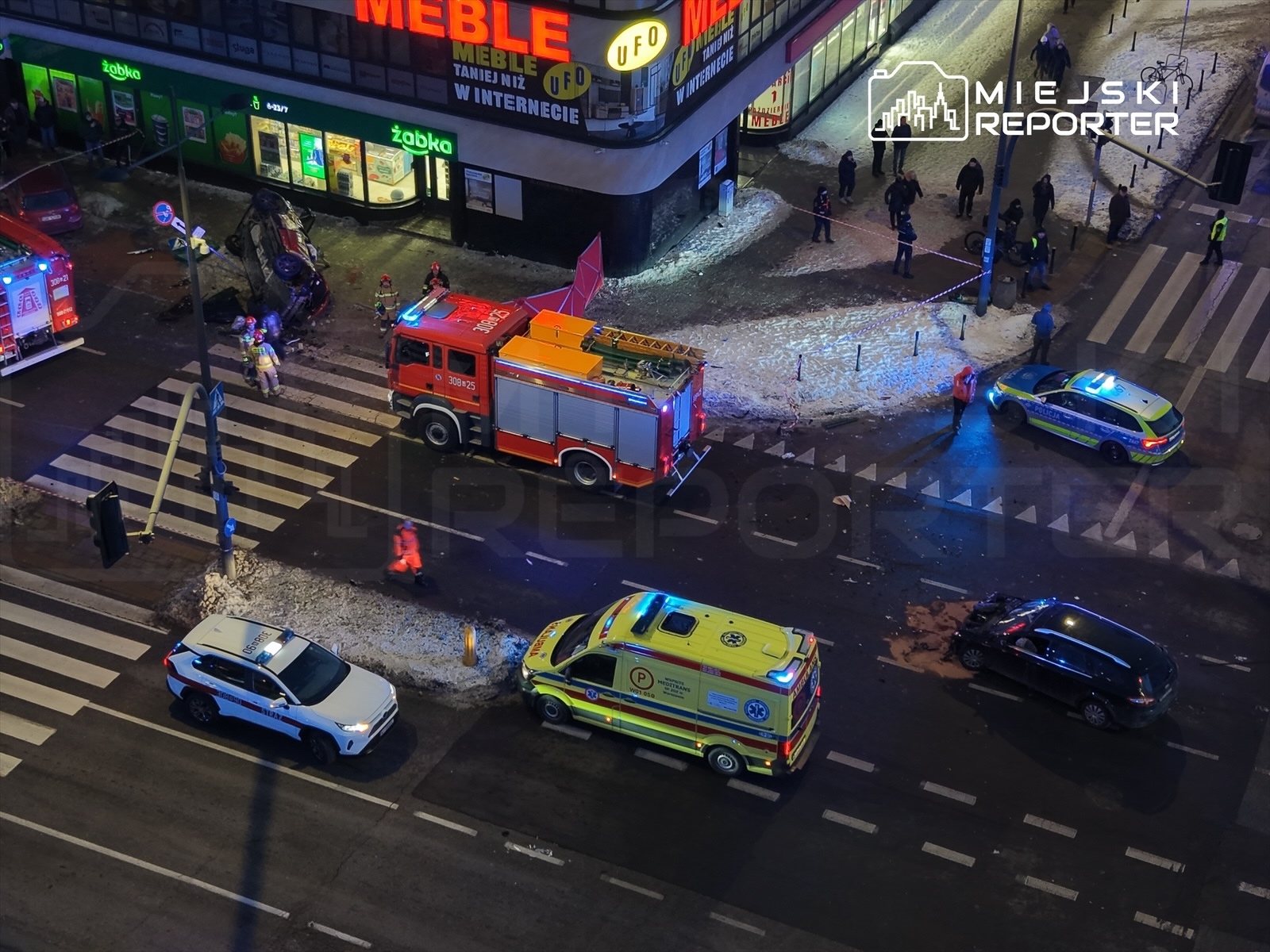 Na skrzyżowaniu, przewrócone auto oraz pojazdy straży pożarnej, policji i karetki, otoczone przez ratowników.