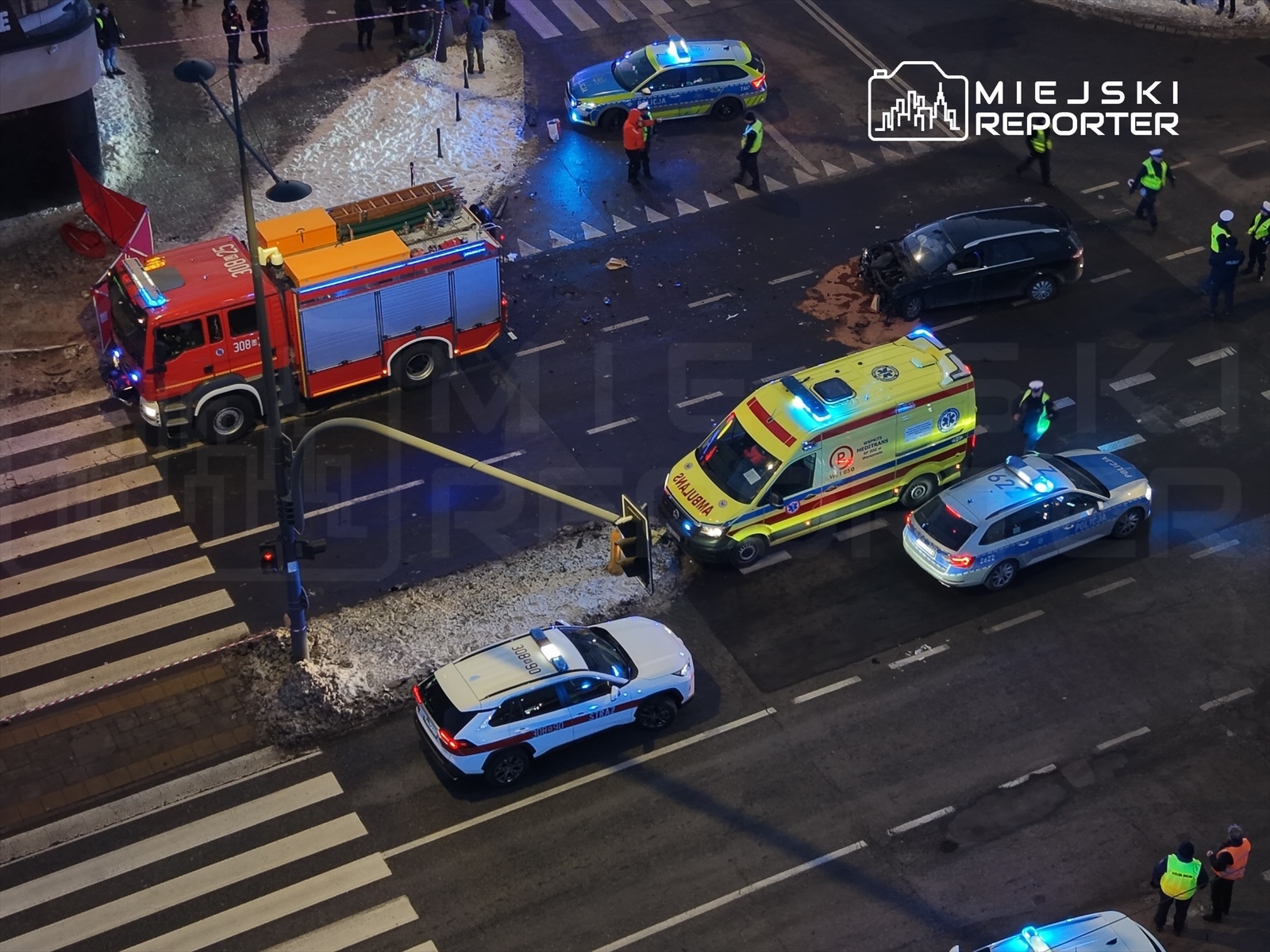 Na skrzyżowaniu stoją woz strażacki, ambulans oraz radiowozy policyjne, a w tle widać ludzi w odblaskowych kamizelkach.