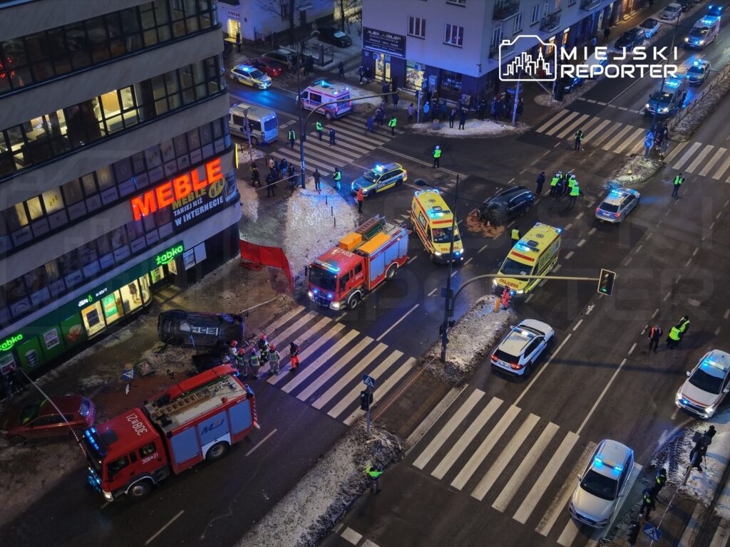 Na skrzyżowaniu ulicy widoczne są służby ratunkowe, w tym karetki i wóz strażacki, obok przewrócony samochód.