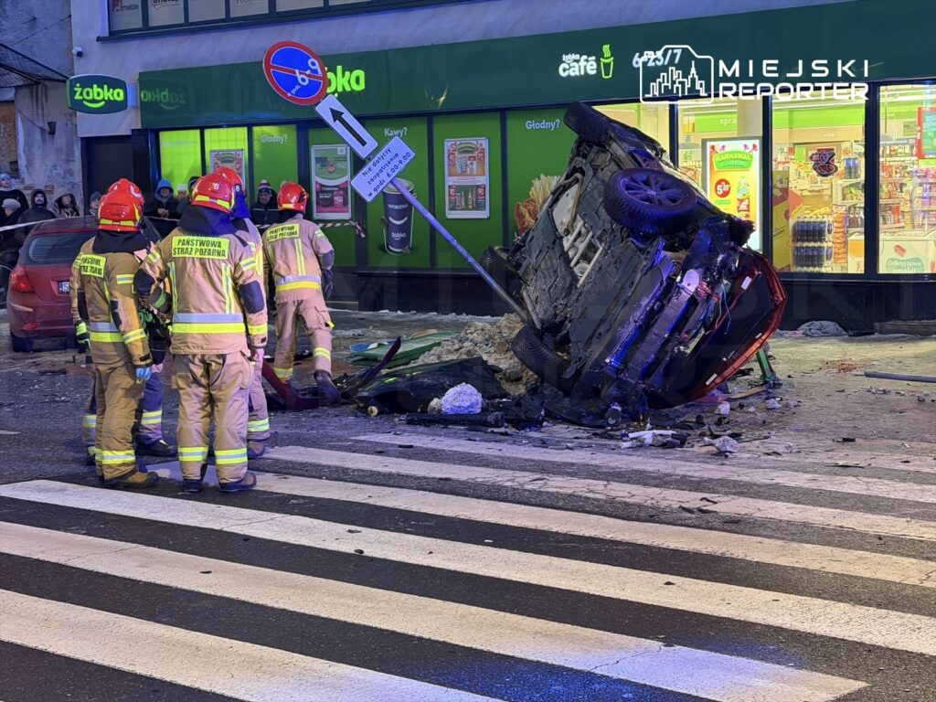 Grupa strażaków w odblaskowych strojach pracuje przy przewróconym samochodzie na ulicy, obok sklepu.