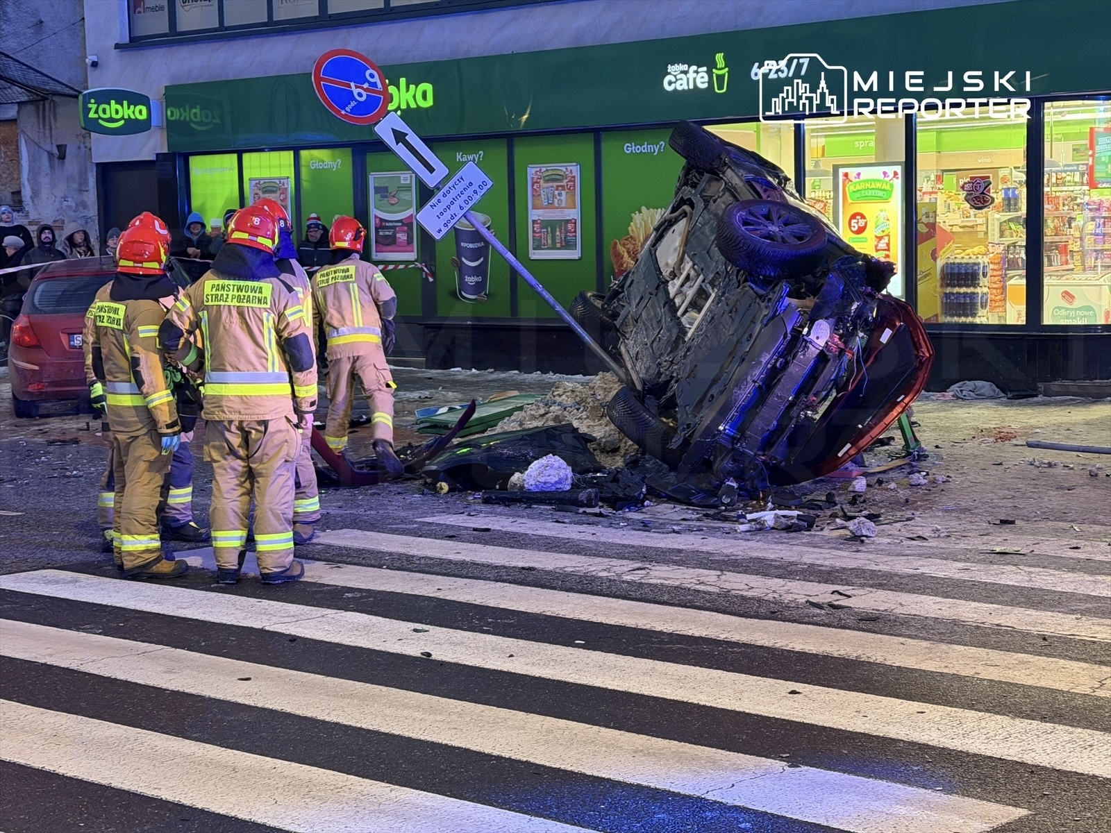 Grupa strażaków w odblaskowych strojach pracuje przy przewróconym samochodzie na ulicy, obok sklepu.