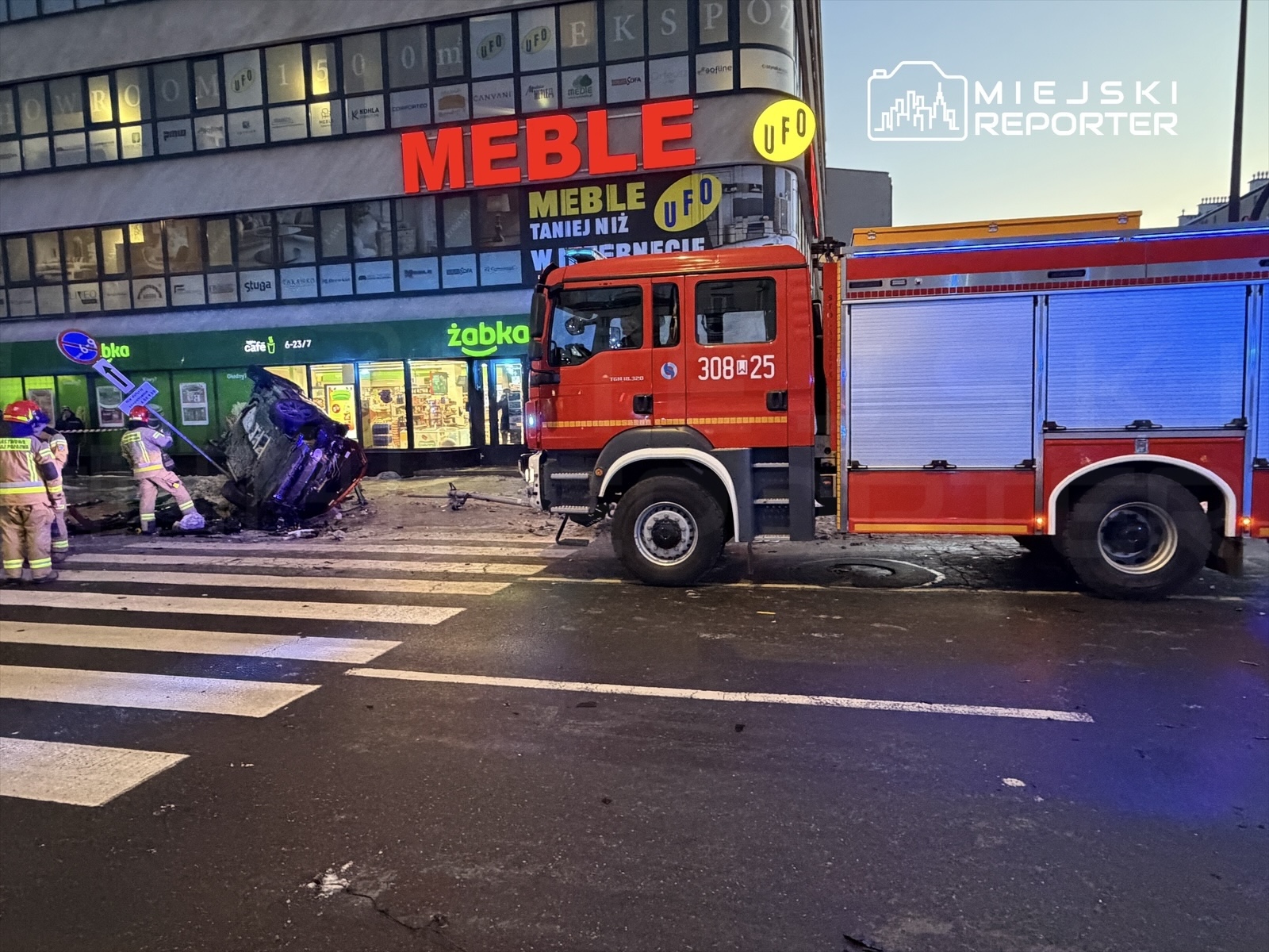 Strażacy pracują nad przewróconym samochodem na skrzyżowaniu, obok sklepu Żabka i biurowca z dużymi oknami.