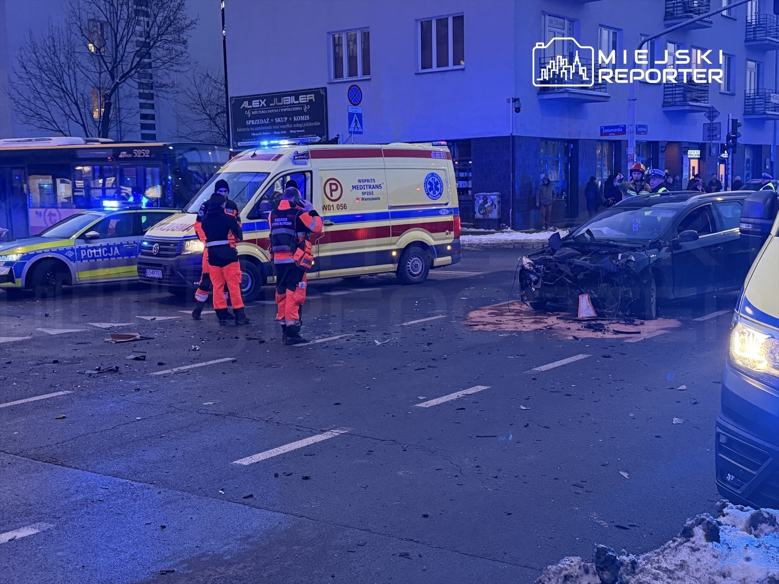 Na skrzyżowaniu ulicy leży uszkodzony samochód, obok stoją ratownicy medyczni i policjanci. W tle widoczne pojazdy służb ratunkowych.