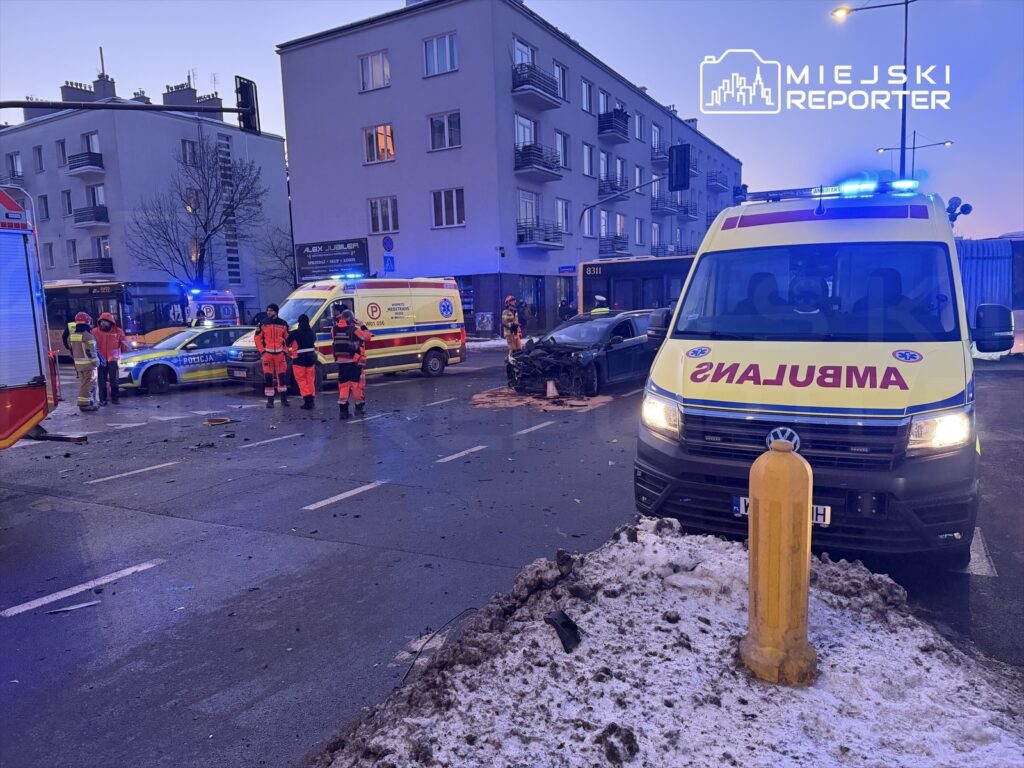 Na skrzyżowaniu ulic pracują ratownicy medyczni i policjanci przy wypadku drogowym, obok uszkodzony samochód i ambulanse.