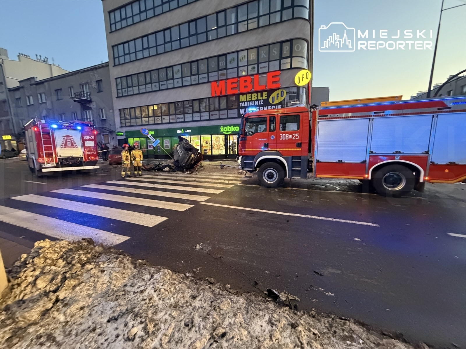 Strażacy zabezpieczają miejsce wypadku, gdzie przewrócone auto leży na jezdni obok nowoczesnego budynku.