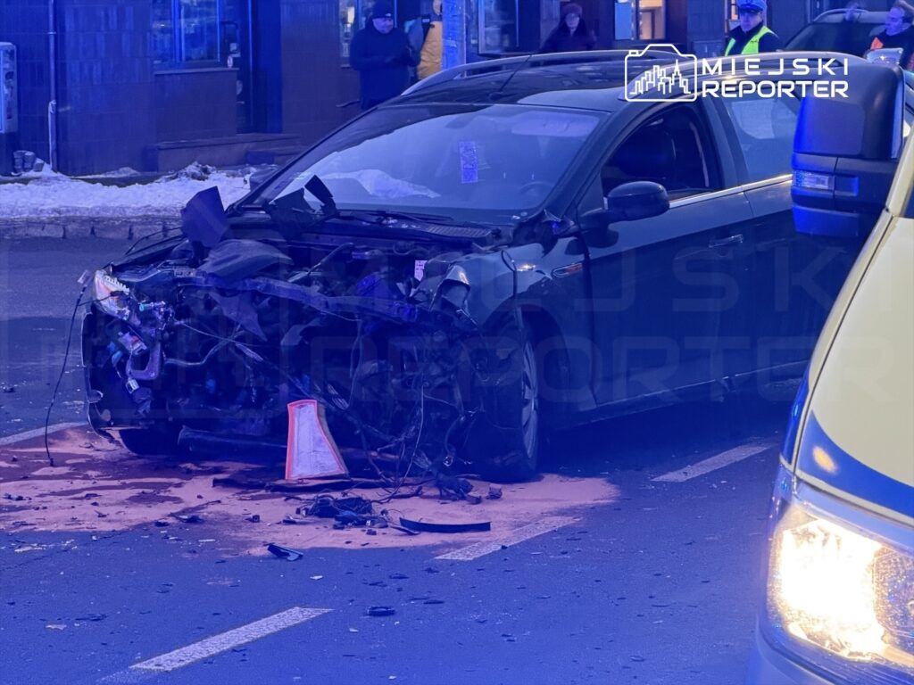 Uszkodzony samochód osobowy z otwartą maską leży na jezdni, obok widać policyjny radiowóz oraz zgromadzonych świadków.