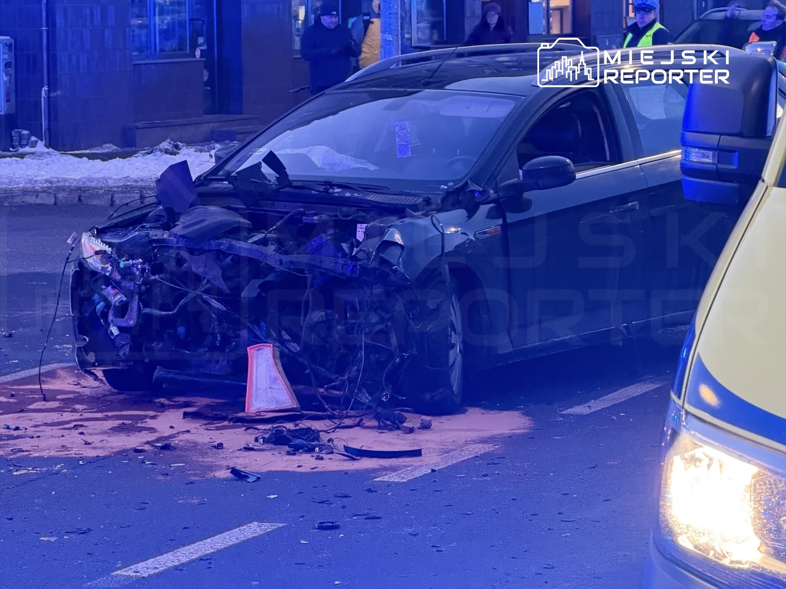 Uszkodzony samochód osobowy z otwartą maską leży na jezdni, obok widać policyjny radiowóz oraz zgromadzonych świadków.