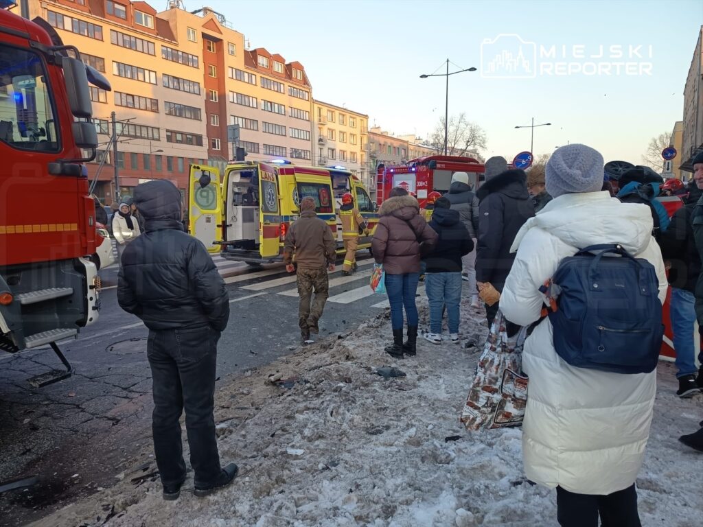 Grupa ludzi obserwująca akcję ratunkową, w tle straż pożarna i karetki, na ulicy pokrytej śniegiem.