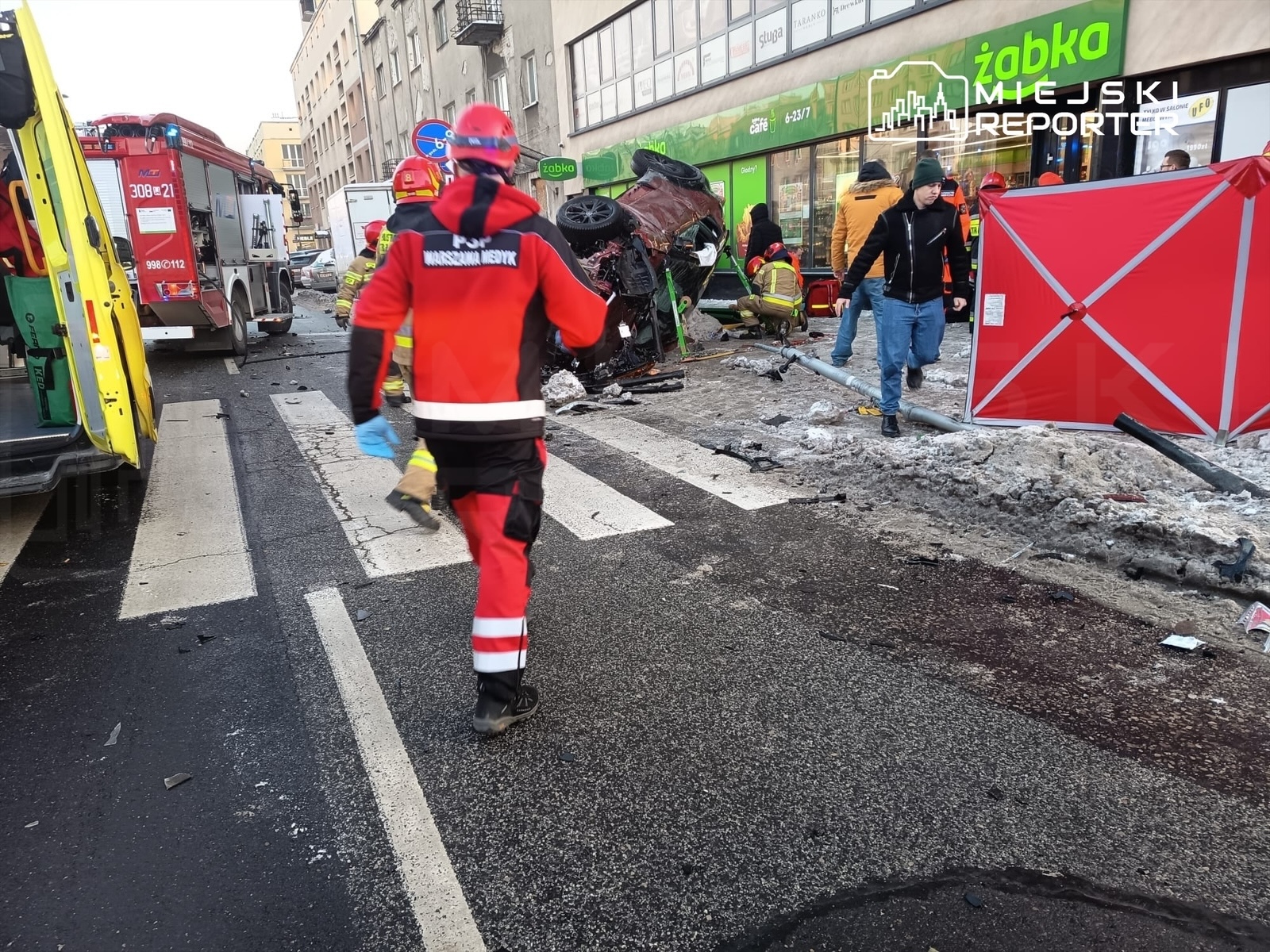 Ratownicy medyczni i strażacy pracują na miejscu wypadku drogowego, w tle przewrócone auto i zniszczenia na jezdni.