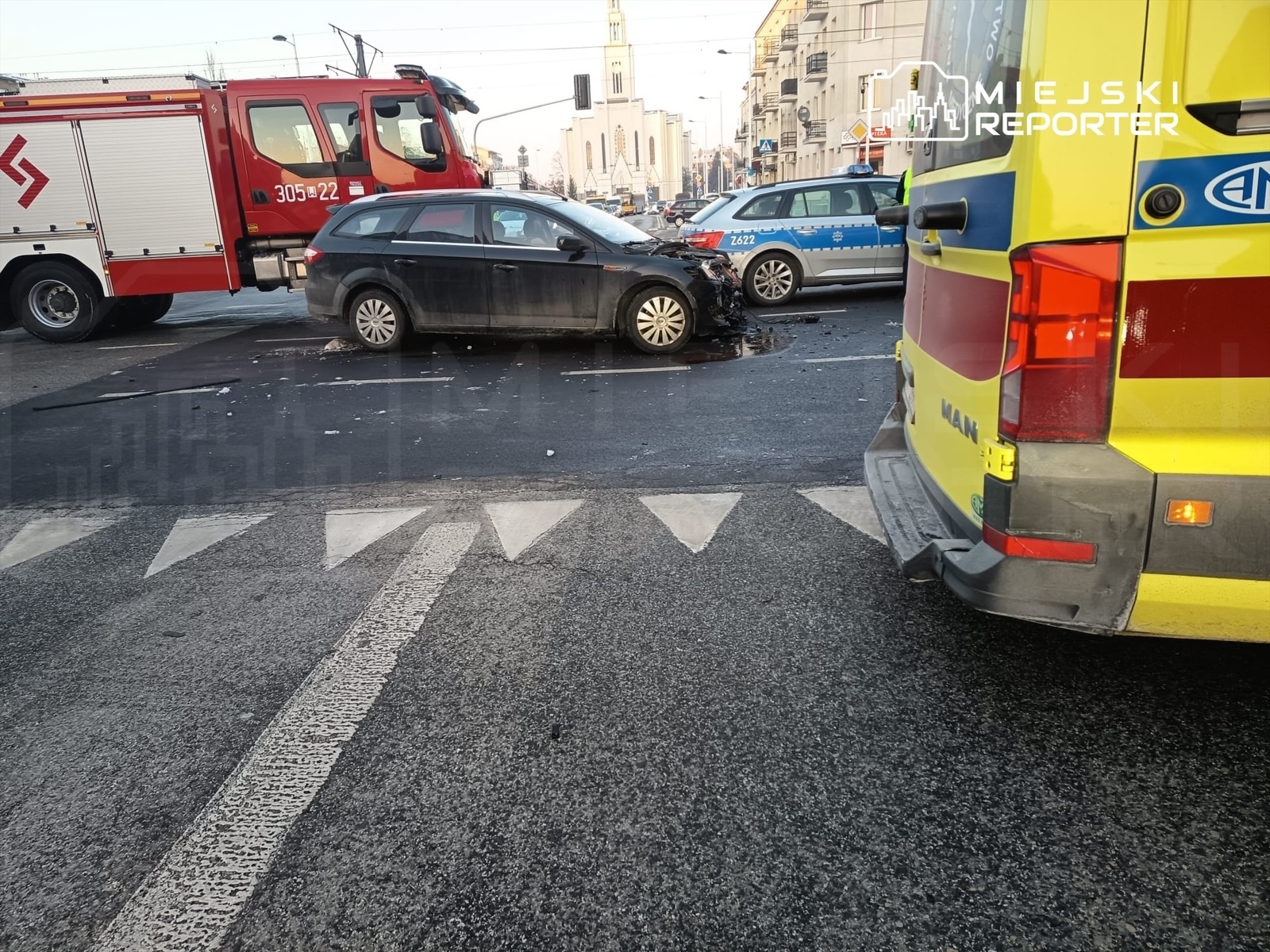 Na skrzyżowaniu stoi uszkodzony samochód osobowy, obok niego widać wóz strażacki i ambulans.
