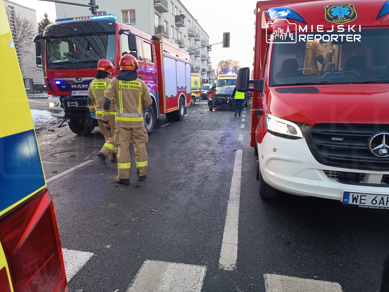 Strażacy w mundurach z żółtymi odblaskami podchodzą do uszkodzonego samochodu na ulicy, obok stoją pojazdy ratunkowe.