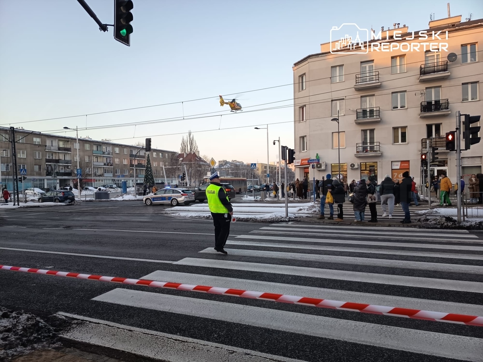 Policjant w żółtej kamizelce kieruje ruchem na skrzyżowaniu, podczas gdy helikopter przelatuje nad miastem.