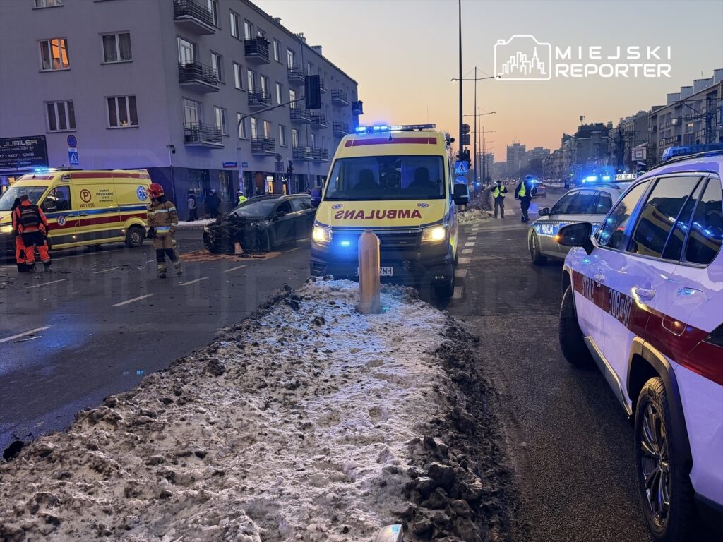 W miejscu wypadku drogowego pracują służby ratunkowe; na jezdni stoi uszkodzony samochód, obok ambulans i radiowóz.
